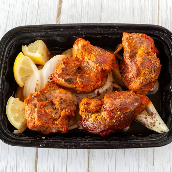 Four pieces of Indian chicken tikka with sliced onions and lemon wedges in a black takeout container.
